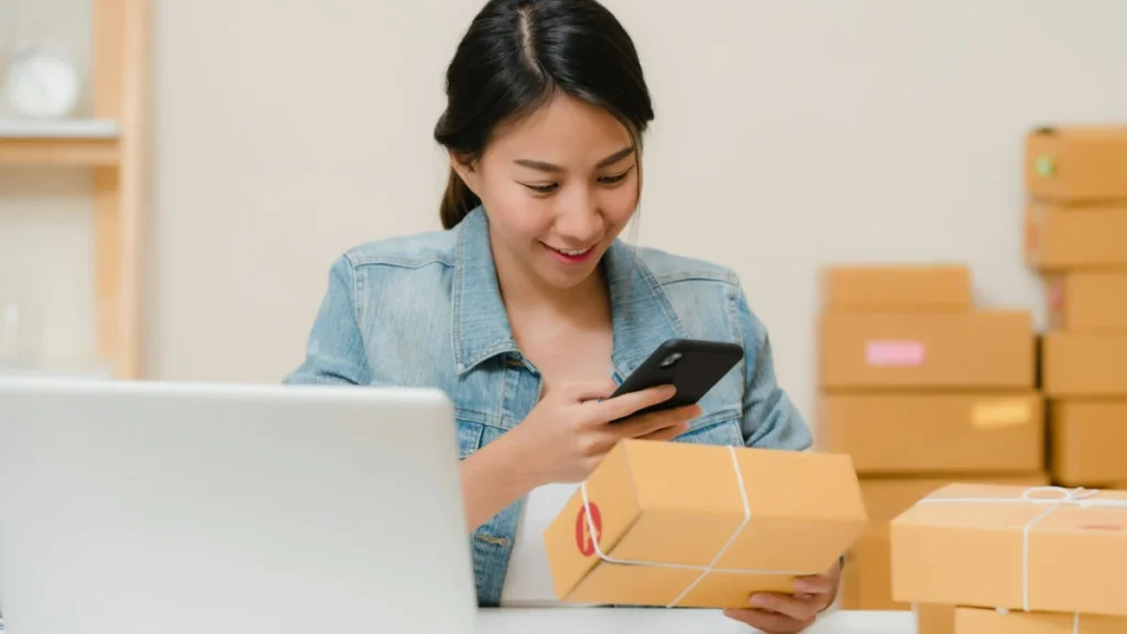 Mulher conferindo um pacote com o celular perto do notebook e de caixas