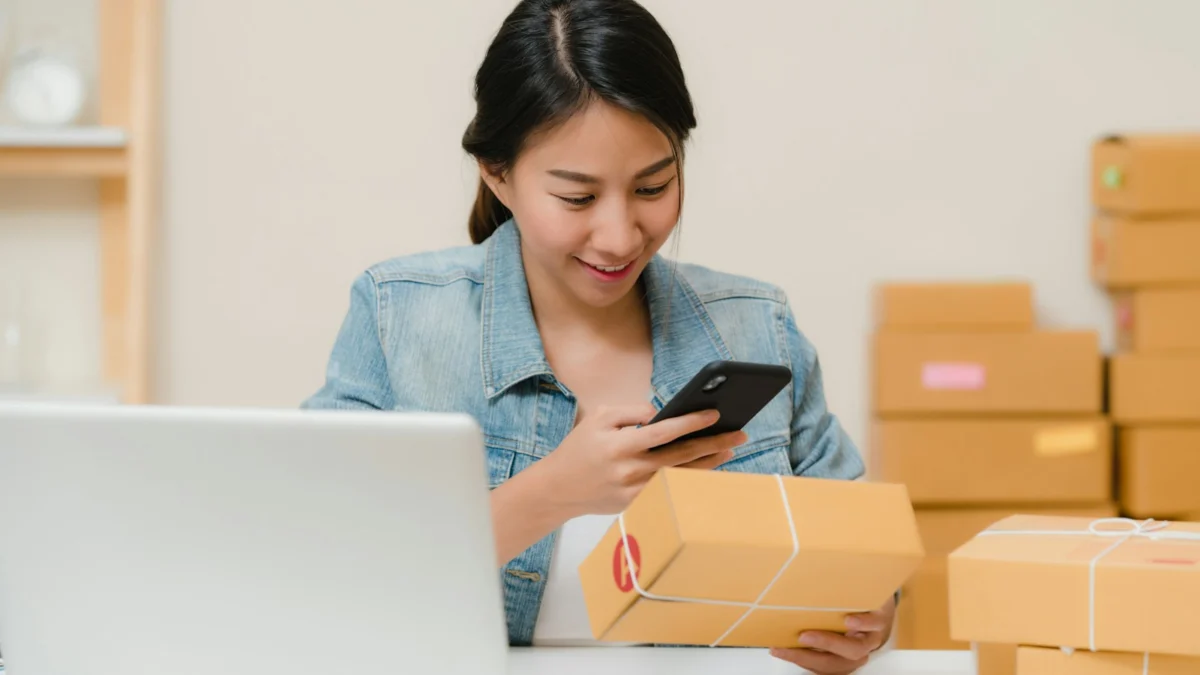 Mulher conferindo um pacote com o celular perto do notebook e de caixas