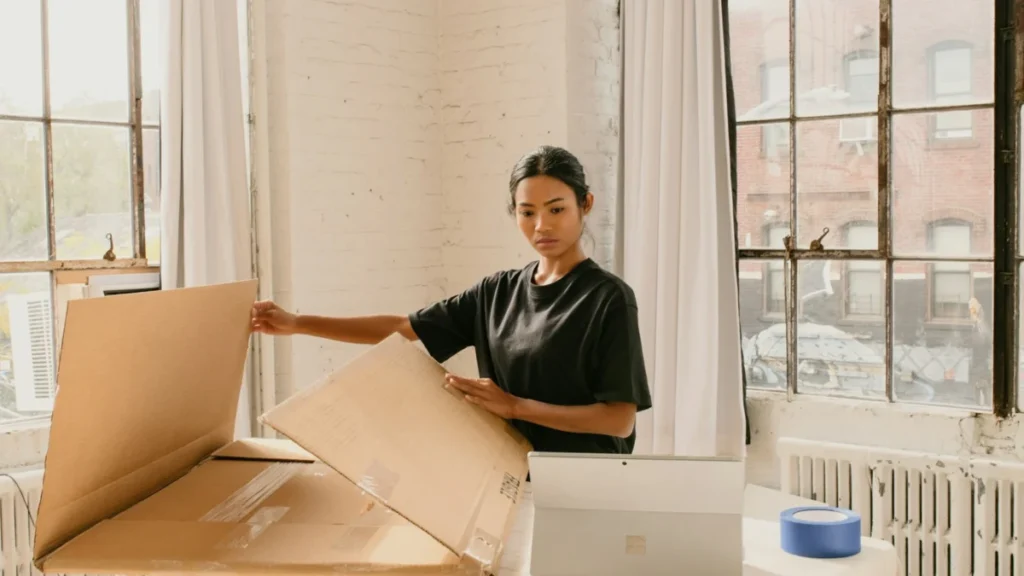 Mulher em pé ao lado de uma caixa de papelão sobre a mesa, preparando um envio